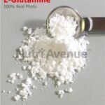 A close-up photograph of white, powdery L-Glutamine supplement piled on a gray surface with a metal measuring spoon scooping some up. The text "L-Glutamine" in red and a "NutriAvenue" watermark are visible.