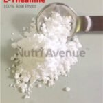 A pile of pure white L-Theanine powder on a flat surface, with a metal measuring spoon scooping a portion of it. The text "L-Theanine," "100% Real Photo," and a "NutriAvenue" watermark are visible on the image.
