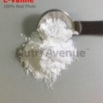 A white, crystalline L-Valine powder piled on a gray surface with a metal measuring spoon scooping a portion of it. Red text in the upper left reads "L-Valine 100% Real Photo," and a translucent watermark for "NutryAvenue" is centered over the powder.